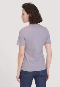 Vrouw met kort bruin haar, gekleed in een lichtpaarse t-shirt met korte mouwen en donkerblauwe jeans, met haar gezicht naar weg van de camera tegen een effen achtergrond.