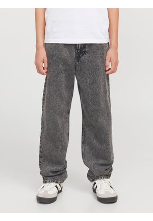 Person iført løstsiddende, falmede sorte jeans, hvide sneakers med sorte striber og en hvid T-shirt, stående mod en ensfarvet baggrund.