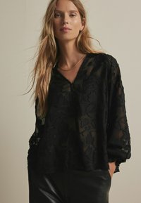 Sort gennemskinnelig bluse med blomsterbroderi, lange ærmer, V-hals og strukturerede detaljer. Bæres med sorte læderbukser.