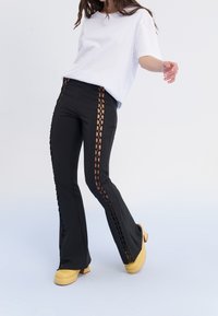 Pantalon noir évasé avec détails lacés sur les côtés, associé à un t-shirt blanc et des chaussures plateforme jaunes. Texture lisse et design moderne.