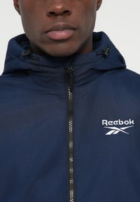 Prim-plan al unei persoane care poartă o jachetă cu fermoar de culoare albastru închis, cu glugă și cu logo-ul alb Reebok pe piept.