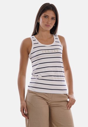 Giovane donna che indossa una canottiera bianca con strisce blu navy e testo, abbinata a pantaloni beige, in piedi davanti a uno sfondo chiaro e semplice.