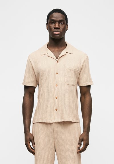 Homme portant une chemise beige à manches courtes avec des boutons et un pantalon assorti au motif chevron subtil, debout devant un fond blanc uni.