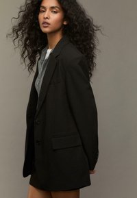 Kvinde med langt krøllet hår iført en sort oversized blazer over en grå lynlås-sweater og en hvid indershirt mod en ensfarvet baggrund.