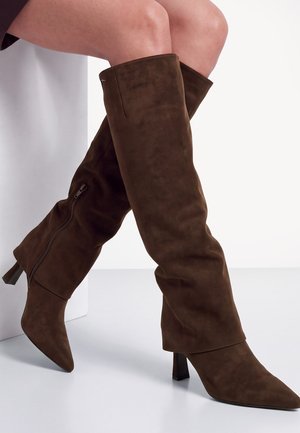 Femme portant des bottes montantes en daim brun foncé avec talons et fermetures éclair latérales, assise jambes croisées sur une surface blanche.