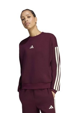 Une femme porte un sweat-shirt Adidas bordeaux à bandes blanches sur les manches et un pantalon de jogging assorti, sur fond blanc.
