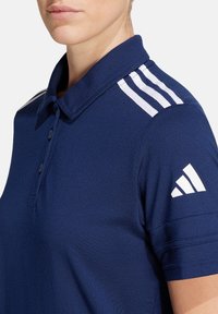 Marineblaues Poloshirt mit klassischem Kragen, das weiße Akzentstreifen in drei Reihen auf den Schultern und das Adidas-Logo auf dem Ärmel aufweist.