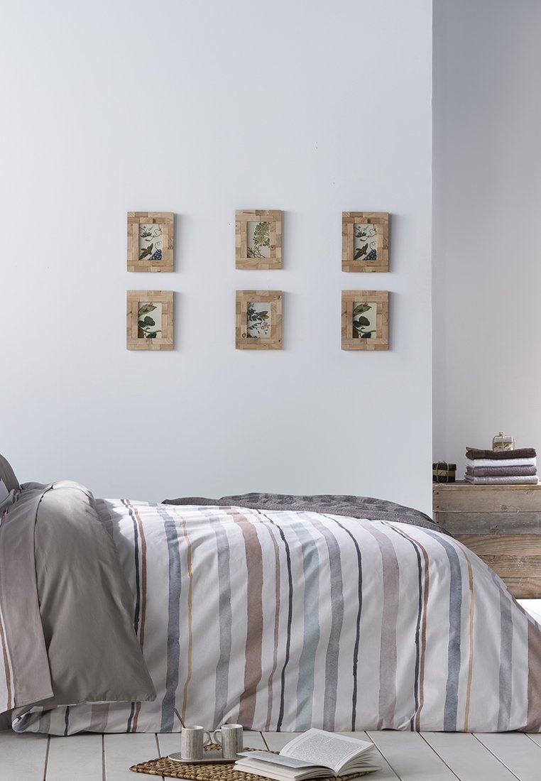 Gestreifte Bettwäsche in Grau-, Beige- und Blautönen, ergänzt durch sechs botanische Drucke in Holzrahmen an einer hellen Wand über einer Holzaufbewahrungsbox.