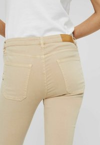 Osoba ubrana w dopasowane beżowe jeansy z tylnymi kieszeniami oraz białą koszulę włożoną do środka, stoi na tle jednolitego jasnego tła.