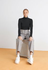 Črn pulover s turtleneck izrezom, prugaste hlače z visokim pasom, bele platforme za športne copate. Model sedi na kovinski škatli z enostavno ozadje.