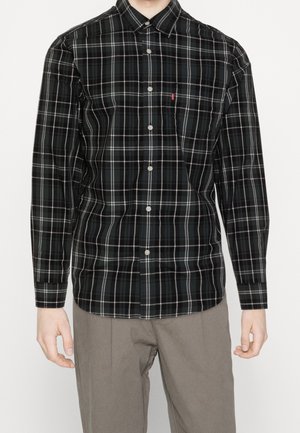 Mann trägt ein schwarz-weiß kariertes Hemd mit langen Ärmeln und hellbraune Hose, steht vor weißem Hintergrund.