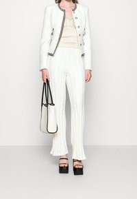 Weißes Tweed-Jacket mit schwarzem Rand, cremefarbenes plissiertes Oberteil, vertikal gestreifte weiße Hose und schwarze Sandalen mit quadratischem Absatz; hält eine Tasche mit schwarzen Akzenten.