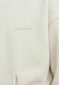 Light beige sweatshirt with a textured fabric, featuring a subtle embossed "ORIGINALS™" logo on the chest and a front pocket.