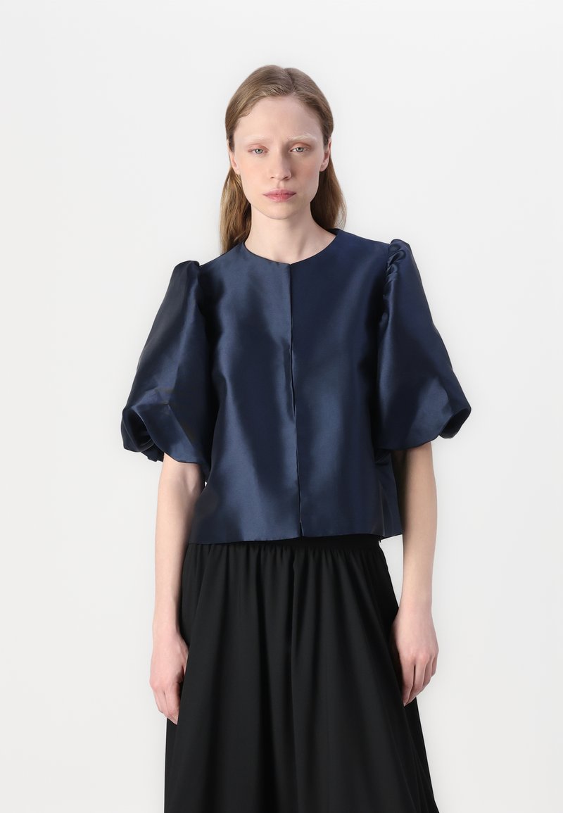 Top corto in satin blu navy con maniche a sbuffo e scollatura rotonda, abbinato a una gonna nera arricciata, creando una silhouette strutturata.