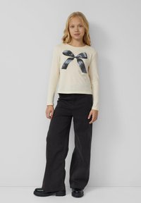 Blondes Mädchen steht und trägt ein cremefarbenes Langarmshirt mit einem schwarzen gepunkteten Schleifenmuster, weite schwarze Hosen und schwarze klobige Schuhe vor einer grauen Wand.