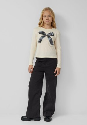 Blond meisje staande, gekleed in een crèmekleurig shirt met lange mouwen en een zwart stippen strikontwerp, brede zwarte broek en zwarte chunky schoenen tegen een grijze muur.