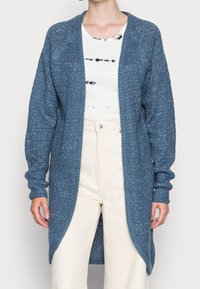 Cárdigan azul de punto con un profundo escote abierto, mangas largas y textura acanalada. Combinado con una camisa blanca y pantalones de color claro.