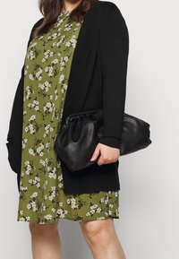 Cardigan nero lavorato a maglia sopra un vestito floreale verde oliva con fiori bianchi. Una borsa a mano in pelle nera è tenuta nella mano destra.