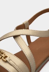 Κοντινό πλάνο μπεζ σανδαλιού με λεπτά δερμάτινα λουράκια και λεπτομέρεια χρυσής αλυσίδας σε καφέ σόλα, με φόντο λευκό.