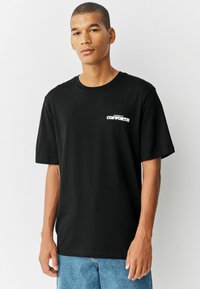 Camiseta de algodón negra con cuello redondo y mangas cortas, que presenta un logo blanco "Powered by COSWORTH" en el lado izquierdo del pecho.