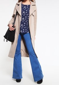 Beige dlouhý kabát, vzorovaný námořnický top s ptáky, modré rozšířené džínsy, černá kabelka na stažení, černá obuv. Hladké textury.