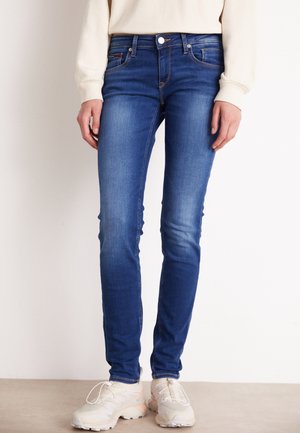 Skinny blue denim jeans with a mid-rise waist, five-pocket design, and subtle fading on the thighs. Paired with light-colored sneakers.