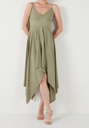 Kobieta ubrana w oliwkowo-zieloną sukienkę bez rękawów z asymetrycznym dołem i beżowe sandały na wysokim obcasie z paskiem wokół kostki, stojąca na tle białej ściany.