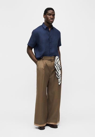 Άνδρας που φοράει μπλε κοντομάνικο πουκάμισο με κουμπιά, καφέ παντελόνι φαρδιάς γραμμής και ύφασμα με ασπρόμαυρο μοτίβο να κρέμεται από την τσέπη.