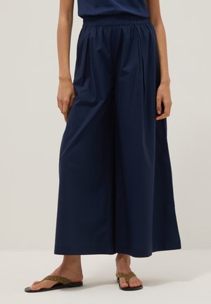 Femme portant un pantalon large bleu marine avec une taille élastique et des sandales plates marron avec des clous circulaires, debout sur un sol clair.