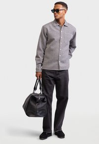 Mann in einem gemusterten Kurzarmhemd, schwarzen Jeans, Sonnenbrille, der eine große schwarze Ledertasche hält und vor einem weißen Hintergrund steht.