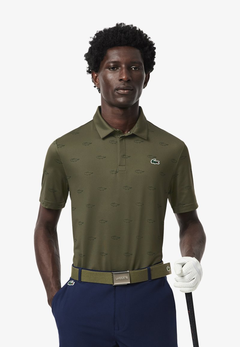 Camisa polo verde con un sutil patrón en toda la prenda, cuello abotonado y logotipo de Lacoste. Combinada con pantalones azul marino y un cinturón a juego.