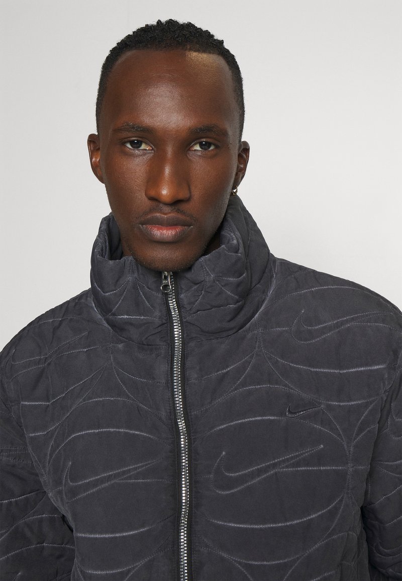 Giacca grigia trapuntata con collo alto, caratterizzata da sottili motivi a spirale testurizzati e chiusura con zip. Logo Nike sul lato sinistro del petto.
