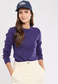 Lila Strickpullover mit langen Ärmeln und Schulterknöpfen, kombiniert mit cremefarbenen Cordhosen. Dunkle Mütze mit weiß gesticktem Logo.
