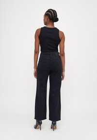 Top cropped preto combinado com jeans pretos de perna larga com discretos brilhos e sapatos de salto alto, visto de costas.