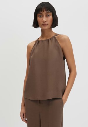 Donna con capelli corti neri che indossa un top marrone senza maniche a collo halter e pantaloni abbinati, in piedi con una mano in tasca.