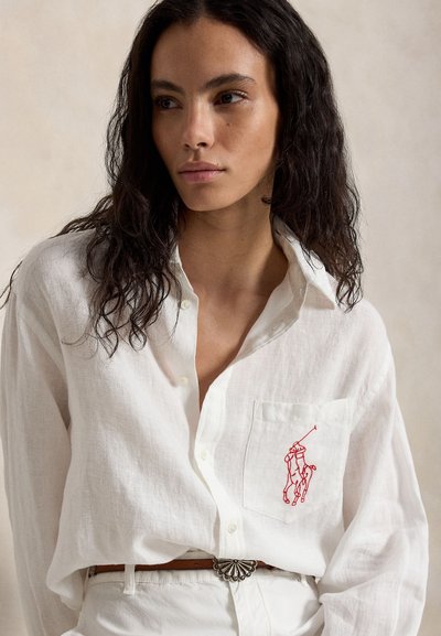 Jongevrouw met lang donker krullend haar draagt een wit overhemd met rode paard en ruiter borduursel op de zak.