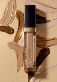 Il correttore Futurist di Estee Lauder in un tubo trasparente con accenti neri e dorati, circondato da vari campioni di crema beige e marrone.