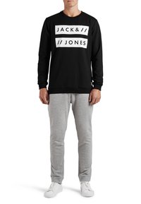 Sudadera negra con logo blanco, cuello redondo y puños de canalé, combinada con pantalones de chándal grises y zapatillas blancas. Ropa deportiva casual.