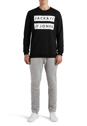 Sweatshirt noir avec logo blanc, col rond et poignets côtelés, associé à un pantalon de jogging gris et des baskets blanches. Vêtements de sport décontractés.
