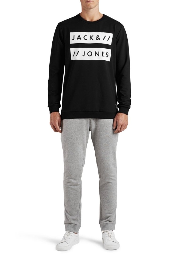 Sudadera negra con logo blanco, cuello redondo y puños de canalé, combinada con pantalones de chándal grises y zapatillas blancas. Ropa deportiva casual.