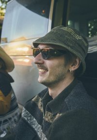 O persoană purtând o pălărie verde închis cu text, ochelari de soare întunecați și o jachetă de lână cu fermoar, multicoloră, zâmbind în aer liber.