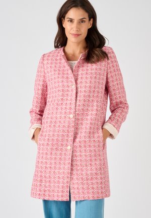 Femme portant un long manteau rose et blanc texturé avec des boutons en perles, les mains dans les poches, debout devant un fond clair uni.