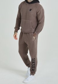 Brun fleecehuva med känguruficka och svart huva; matchande sweatpants med logotypdetalj på vänster ben och elastisk midja; vita sneakers.
