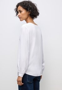 Blusa bianca a maniche lunghe con una vestibilità rilassata, tessuto morbido, scollo rotondo e polsini con bottone, abbinata a jeans in denim blu scuro.