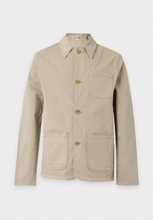 Homme portant une veste beige boutonnée sur une chemise blanche, jean bleu ample et chaussures marron, debout devant un fond uni.