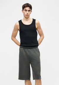 Jeune homme portant un débardeur noir sans manches et un short gris mi-genou, debout les mains derrière le dos sur un fond blanc.