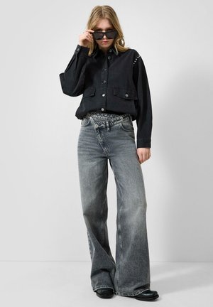 Junge Frau trägt eine schwarze, knöpfbare Jacke, weite ausgewaschene graue Jeans, schwarze Schuhe und richtet ihre schwarze Sonnenbrille vor einem einfarbigen Hintergrund.