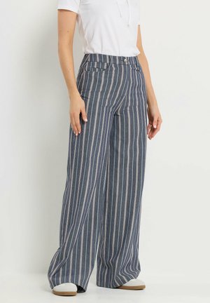 Donna che indossa pantaloni a vita alta, ampi e blu con righe verticali bianche, abbinati a una maglietta bianca a maniche corte e scarpe da ginnastica bianche.