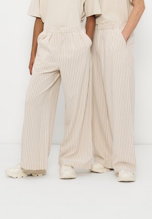 Deux personnes portant des pantalons beiges à larges jambes à fines rayures avec des ceintures élastiques et des baskets blanches, debout côte à côte.