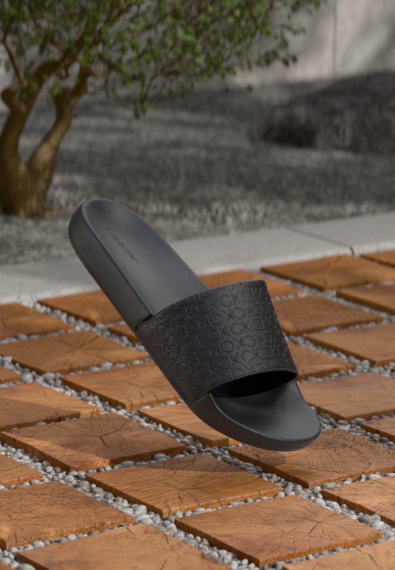 Infradito neri Calvin Klein appoggiati su un pavimento in piastrelle di legno con piccole pietre grigie, sfondo sfocato di un albero e un muro di cemento.